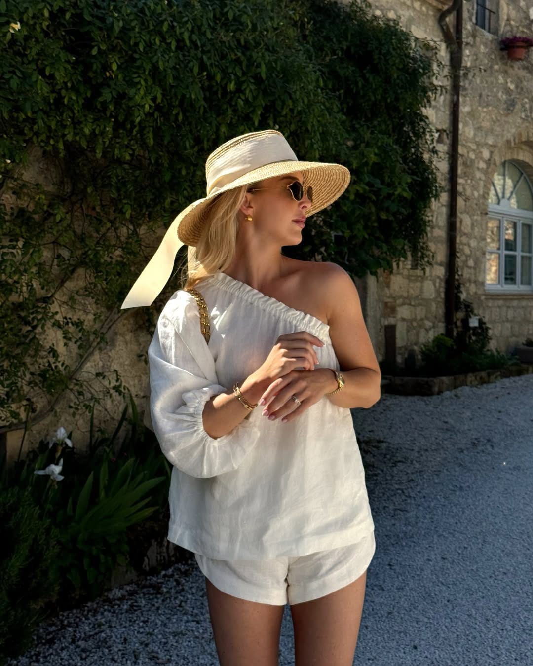 Woman in white linen one-shoulder top and shorts with straw ribbon hat and gold chain bag outdoors.