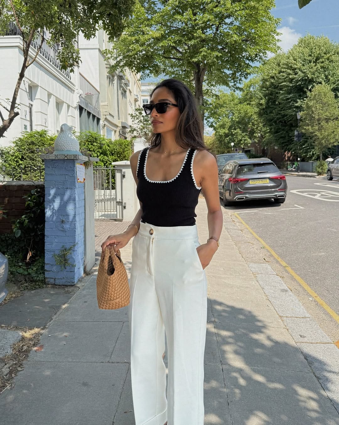 Woman in black scallop-trim ribbed tank and white wide-leg tailored trousers with woven tan bucket bag on a street.