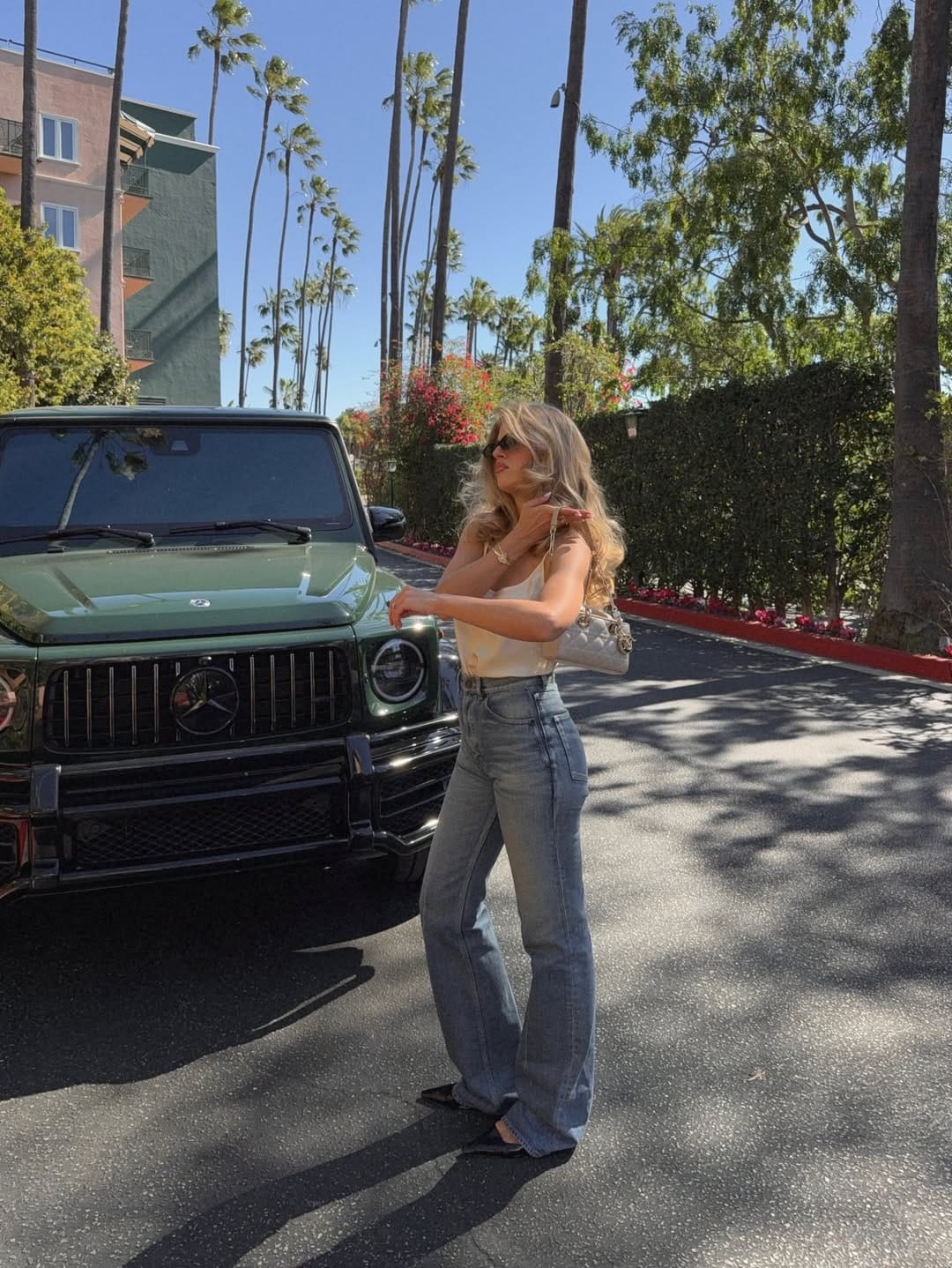 Woman in cream silk cami and wide-leg mid-wash blue jeans with black pointed flat mules and quilted chain bag.