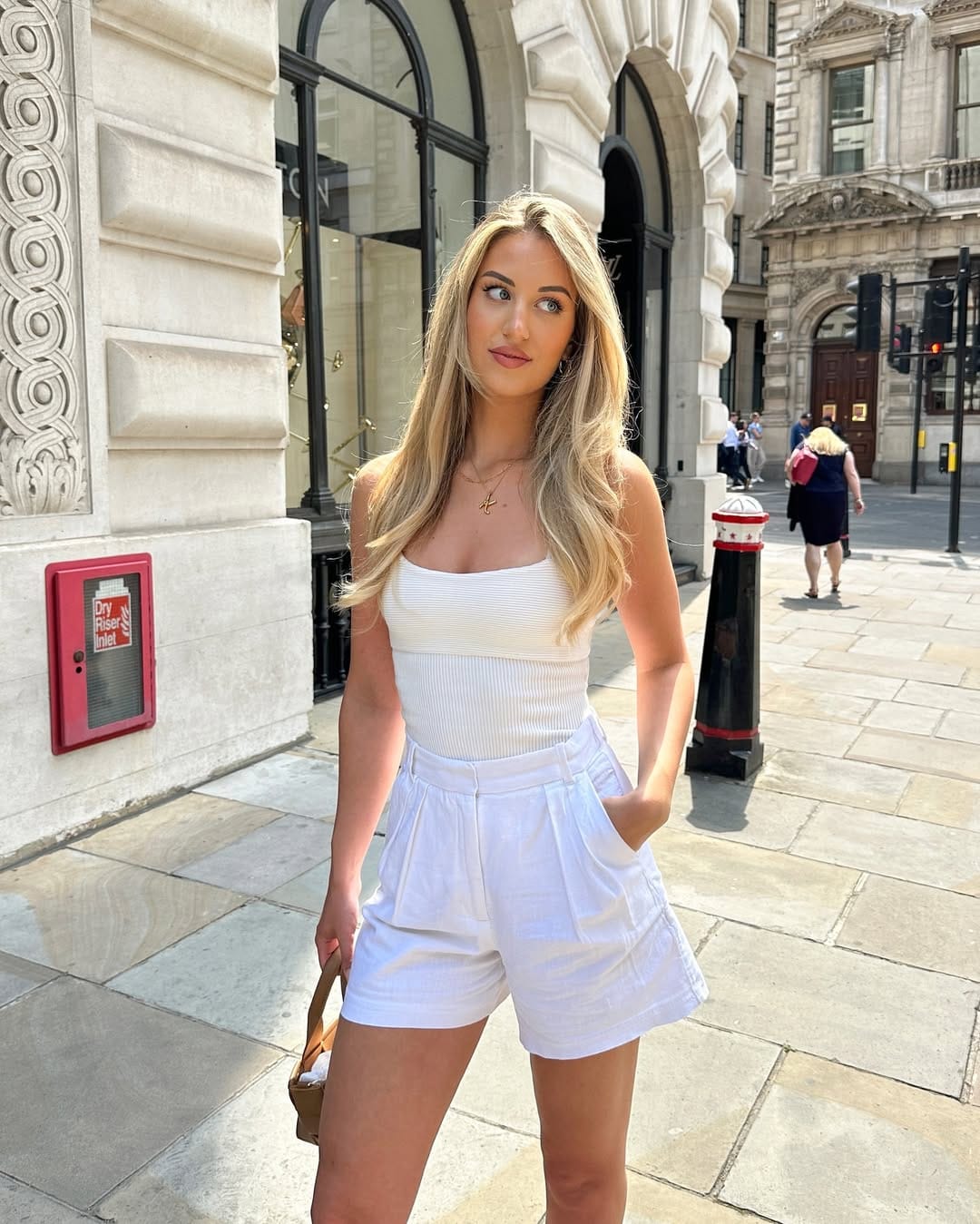 Woman in white strapless ribbed bodysuit and white pleated linen shorts with gold necklace on a city street.