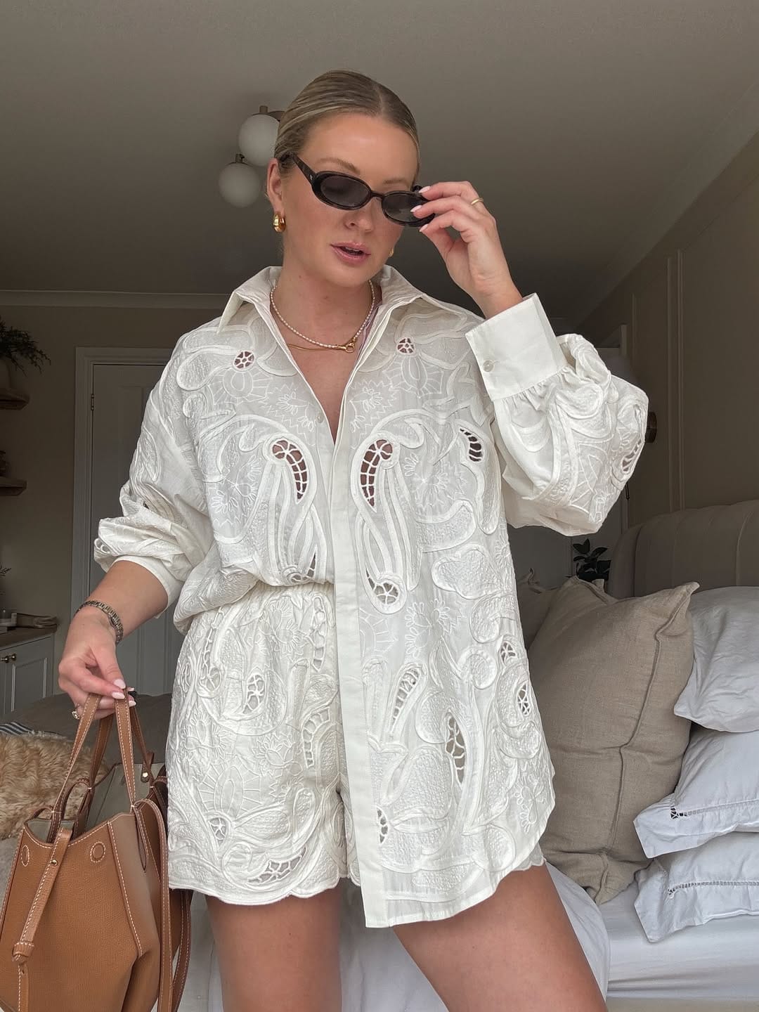 Woman in white cutwork embroidered oversized shirt and matching shorts with pearl necklace and tan leather tote.
