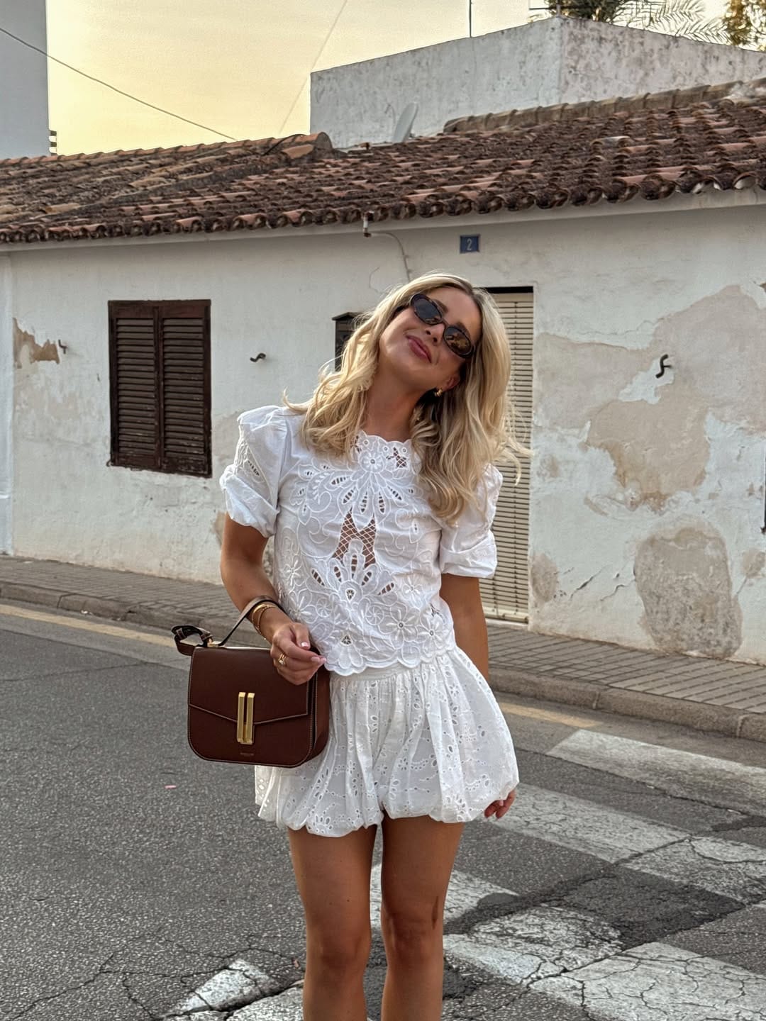 Woman in white broderie anglaise puff-sleeve top and scalloped mini skirt with brown saddlebag on a street.