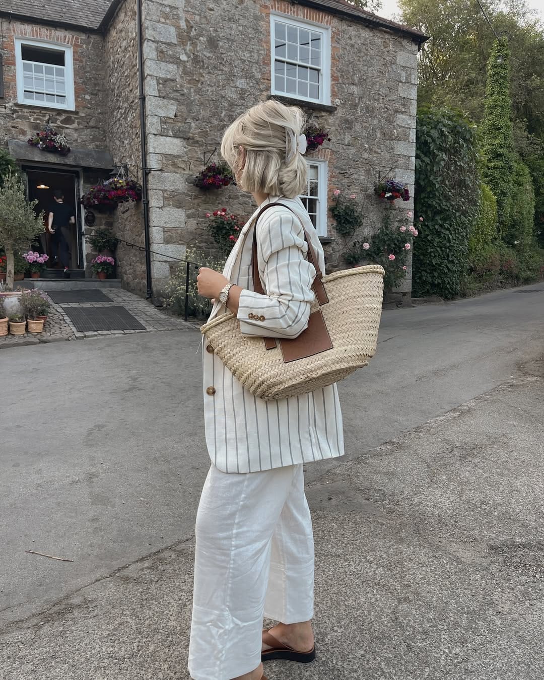 Woman in cream pinstripe longline linen blazer, white linen trousers and large woven straw basket tote bag.
