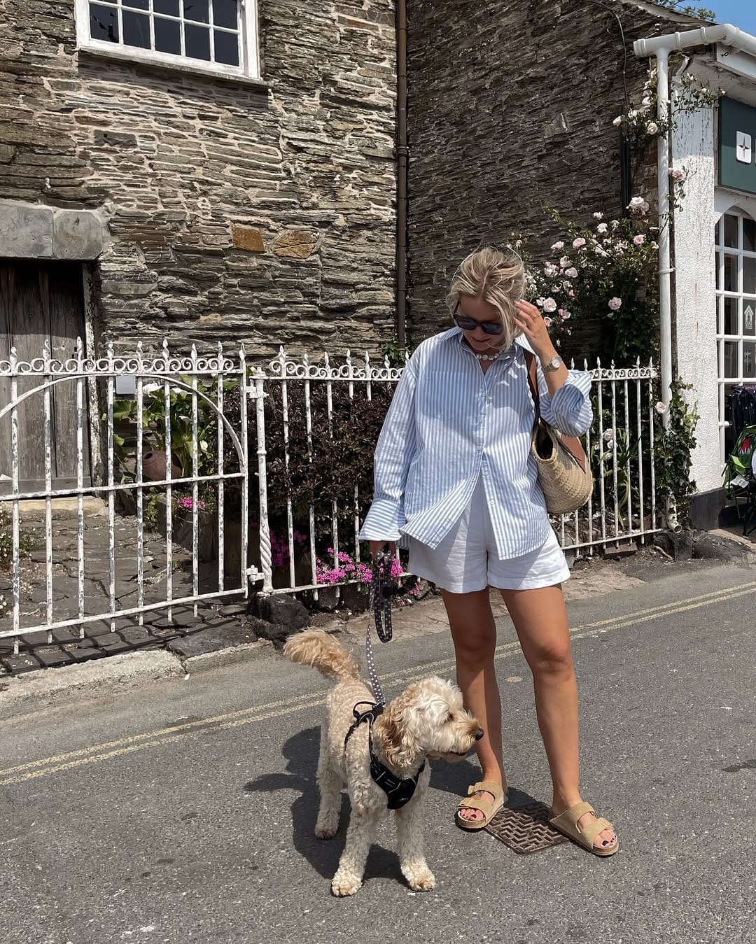 Woman in blue stripe oversized shirt, white linen shorts and tan flat sandals walking with a small dog.