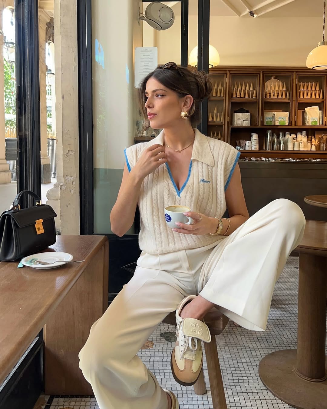 Woman in cream cable-knit polo vest, ivory wide-leg trousers and cream Adidas sneakers seated in a café.
