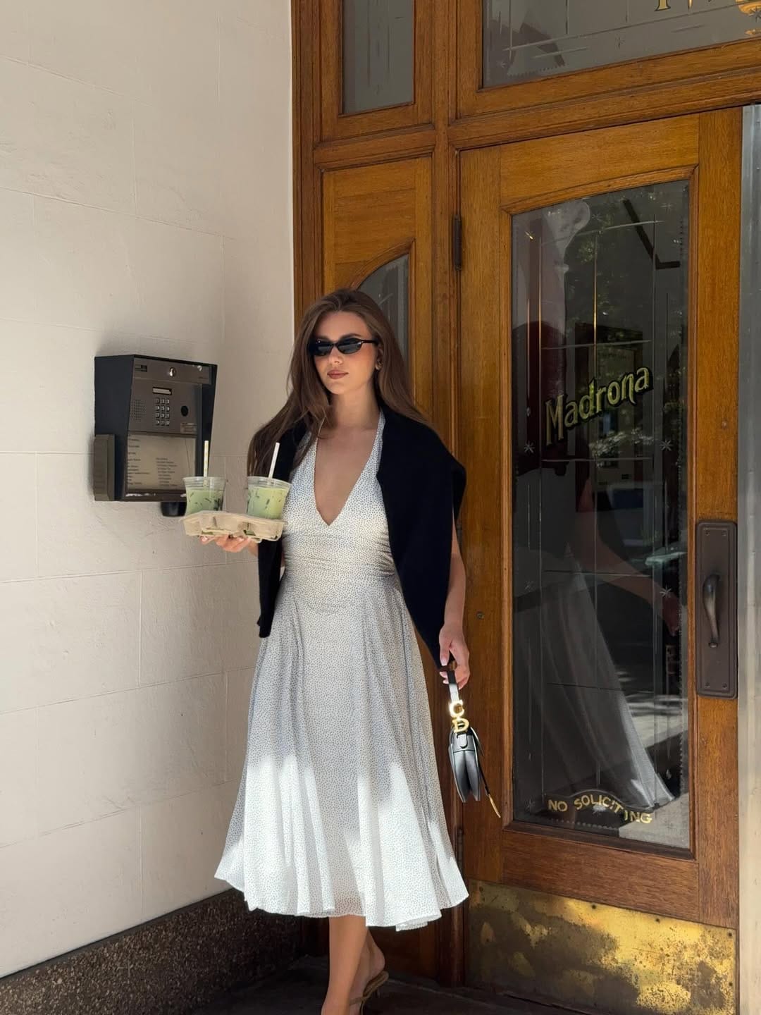 Woman in white spot midi dress with black short-sleeve cardigan draped over shoulders and black mini bag.