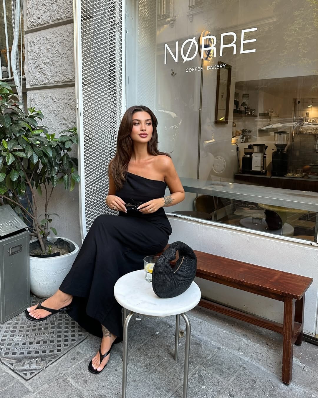 Woman in all-black one-shoulder wide-leg jumpsuit with woven black bag seated outside a café.