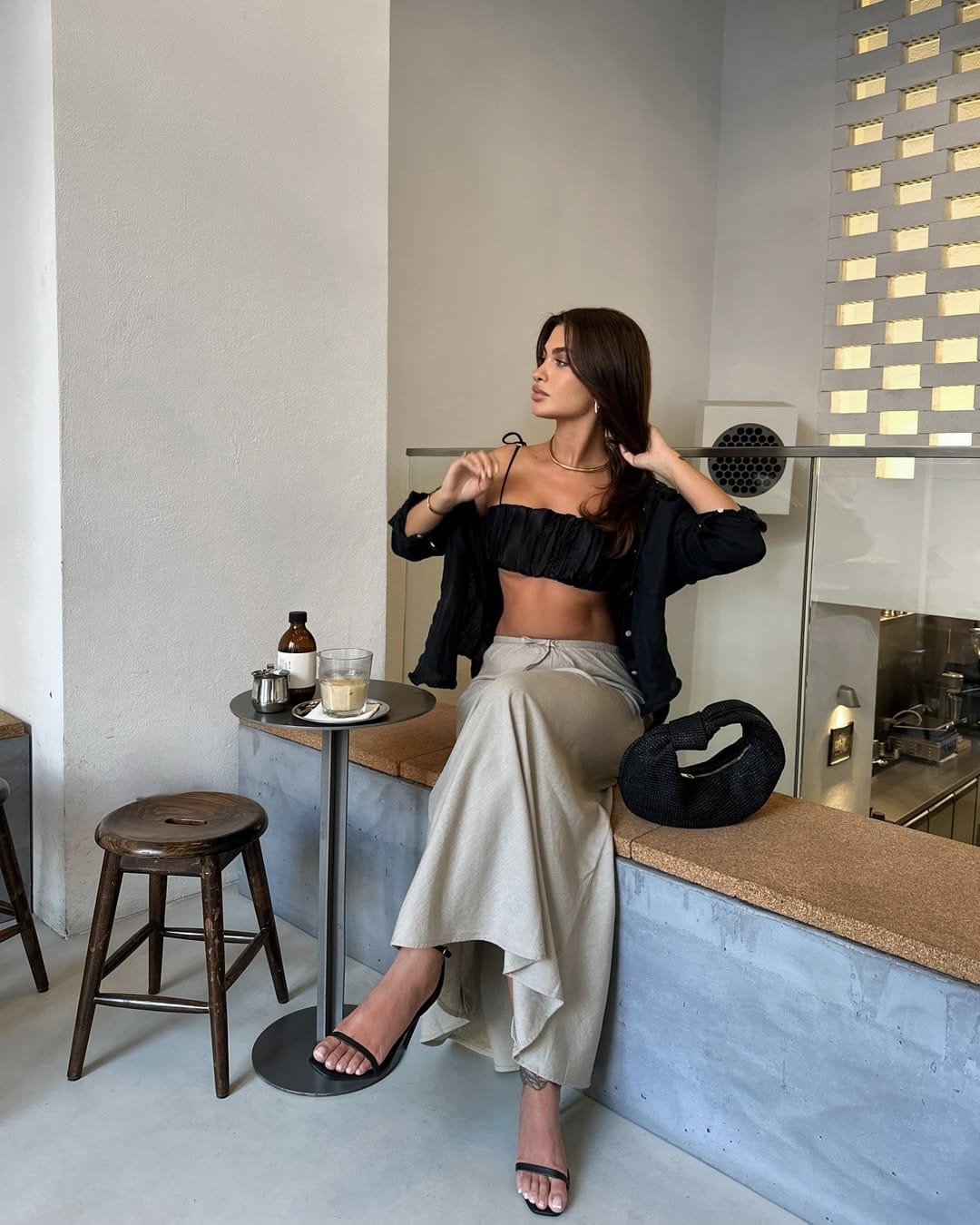 Woman in black ruched crop top, open overshirt and stone wide-leg linen trousers with black heeled sandals in a café.