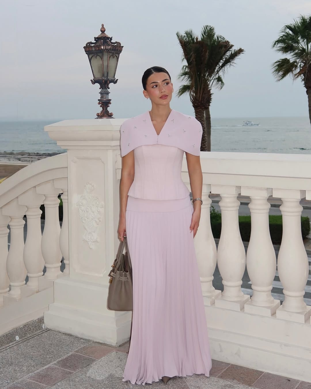 Woman in blush pink structured cape peplum top and pleated maxi skirt with taupe bag by the sea.
