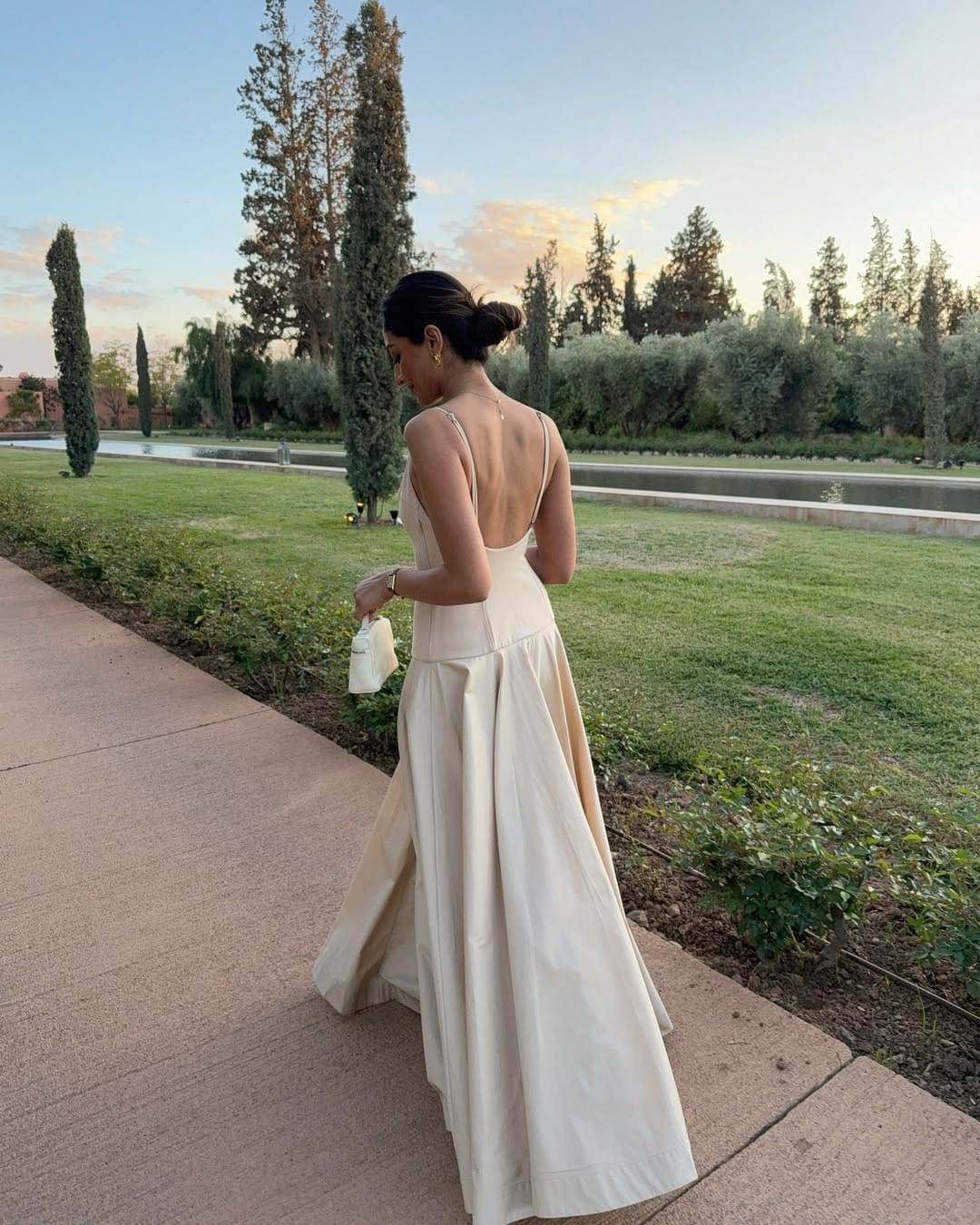 Woman in champagne satin open-back spaghetti-strap ball gown with white clutch at sunset in a Moroccan garden.