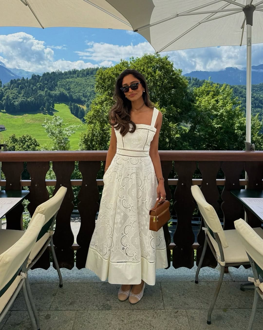 Woman in white broderie anglaise square-neck structured midi dress with tan vanity bag on an alpine terrace.