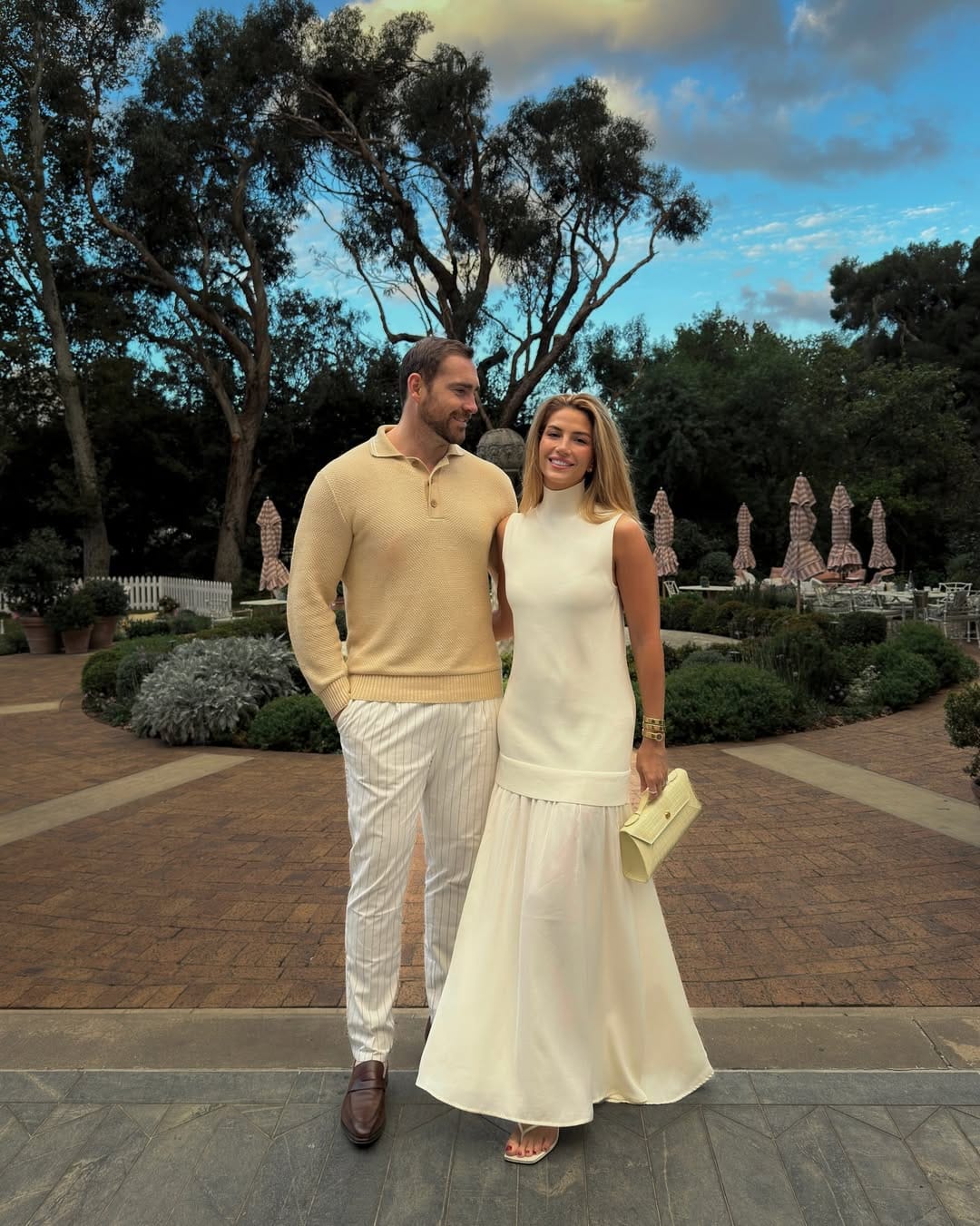 Woman in ivory sleeveless mock-turtleneck drop-waist maxi dress with yellow clutch at a garden wedding.