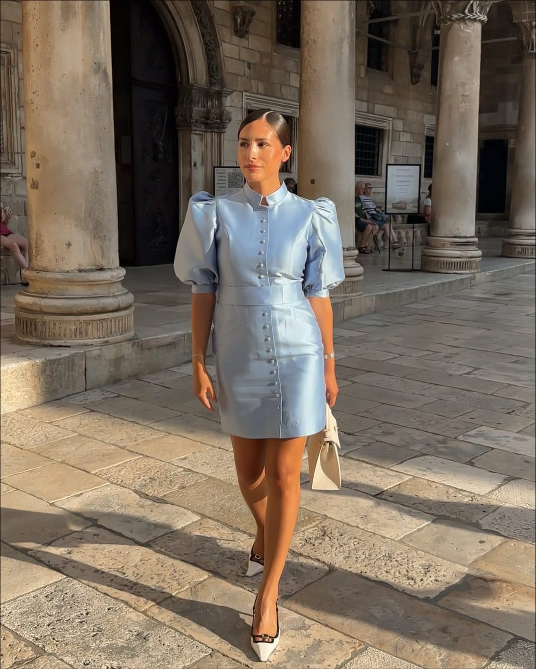 Woman in baby blue puff-sleeve button-front satin mini dress with white slingback heels in Dubrovnik.