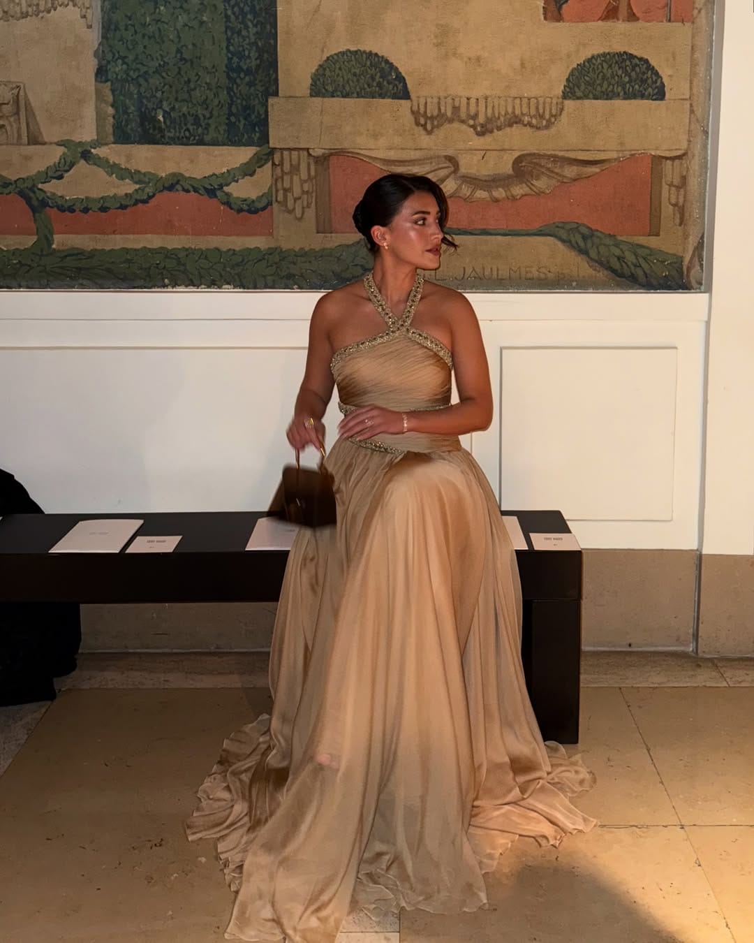 Woman in champagne gold cross-halter pleated ballgown with gold beaded neckline inside a frescoed interior.