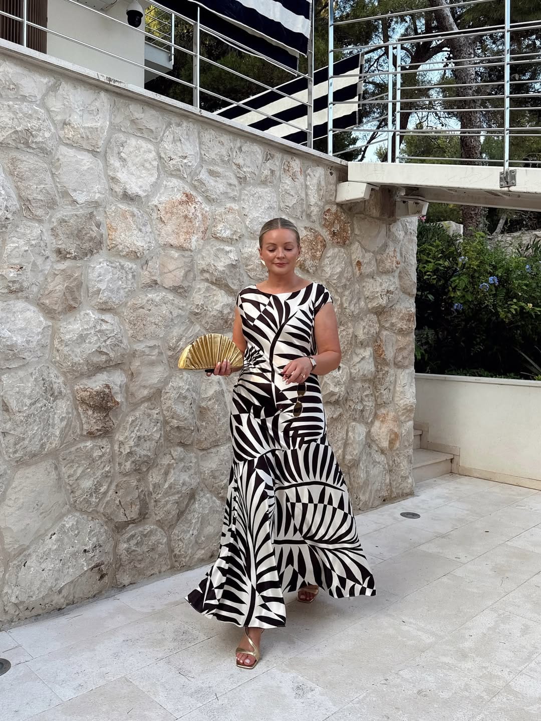 Woman in black white abstract print cap-sleeve maxi dress with gold fan clutch and gold sandals at a Croatian venue.