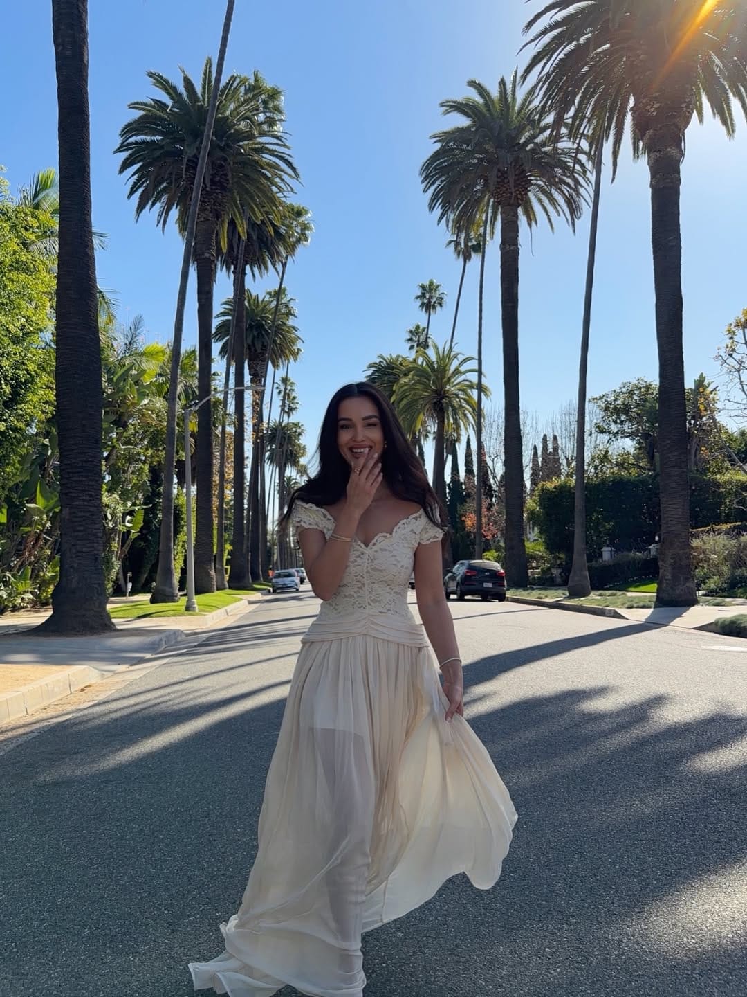 Woman in ivory off-shoulder lace bodice chiffon ball gown twirling on a sunny palm-lined boulevard.