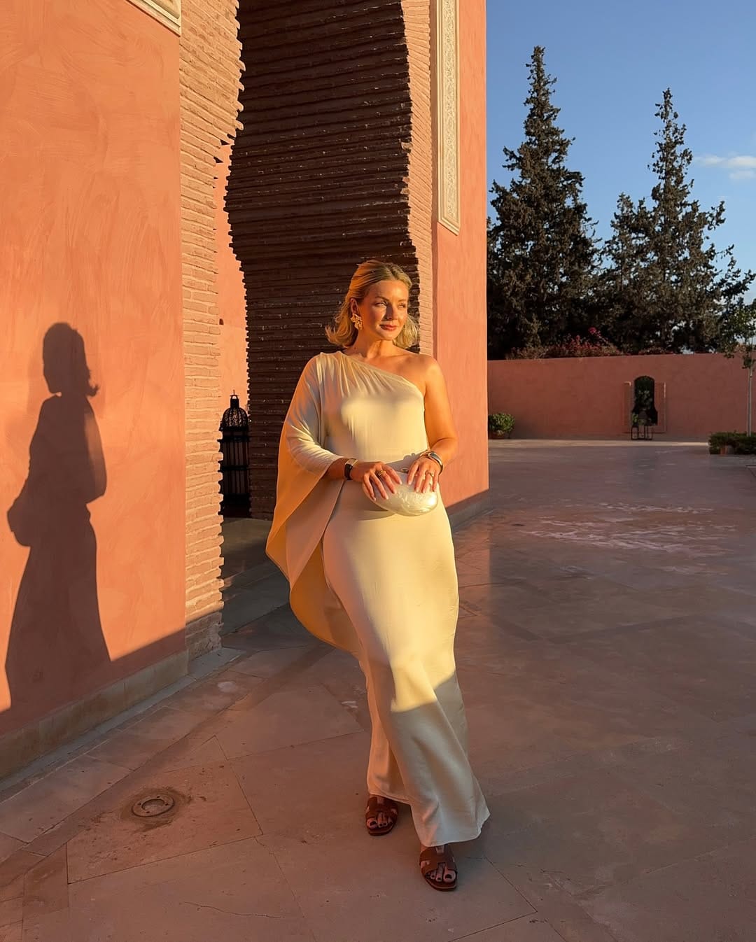 Woman in ivory one-shoulder cape maxi dress with white clutch and brown Hermès sandals in a Moroccan riad courtyard.