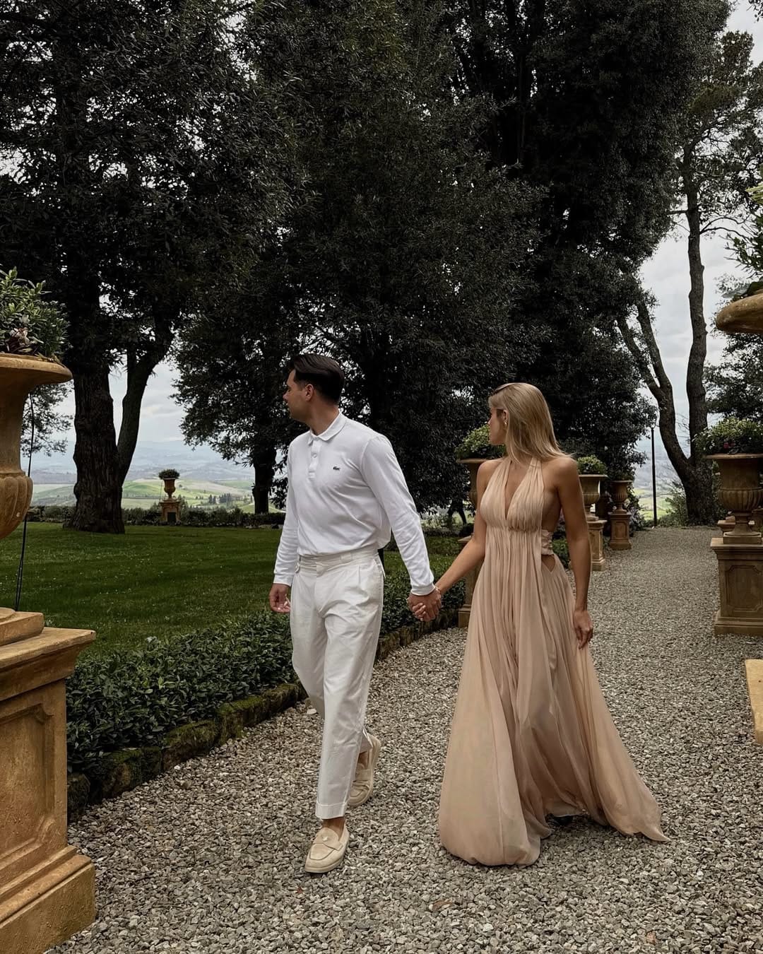 Woman in blush nude pleated halterneck maxi gown walking on a gravel avenue at a Tuscan villa wedding.