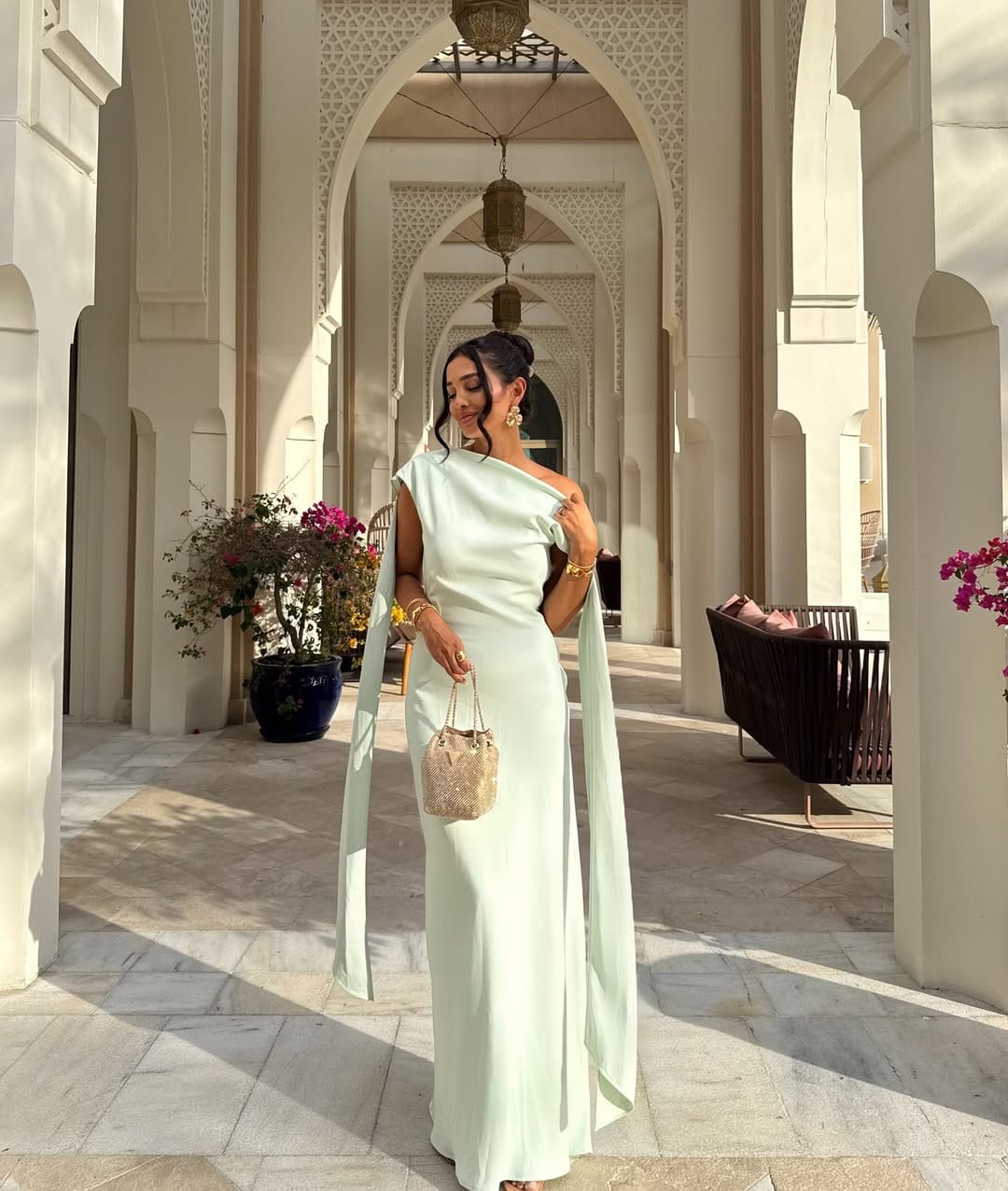 Woman in mint green one-shoulder cape sleeve column gown with gold bag inside a Moorish arched corridor.