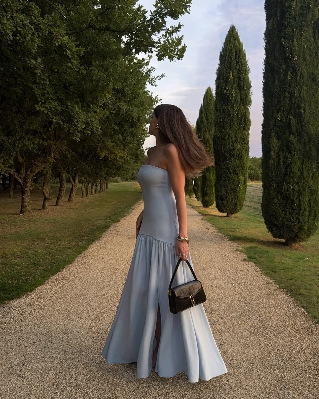 Woman in powder blue strapless drop-waist maxi gown with black top-handle bag on a cypress-lined path in France.