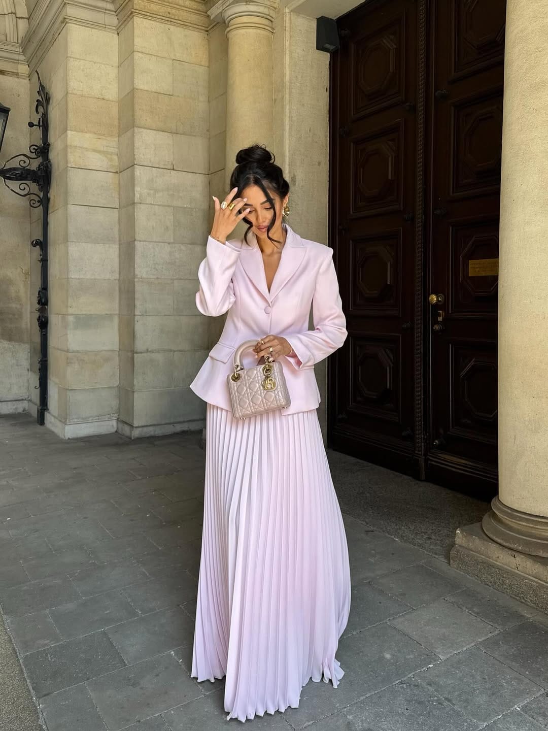 Woman in lilac satin blazer and matching accordion-pleated maxi skirt with pink Lady Dior bag outside a stone building.
