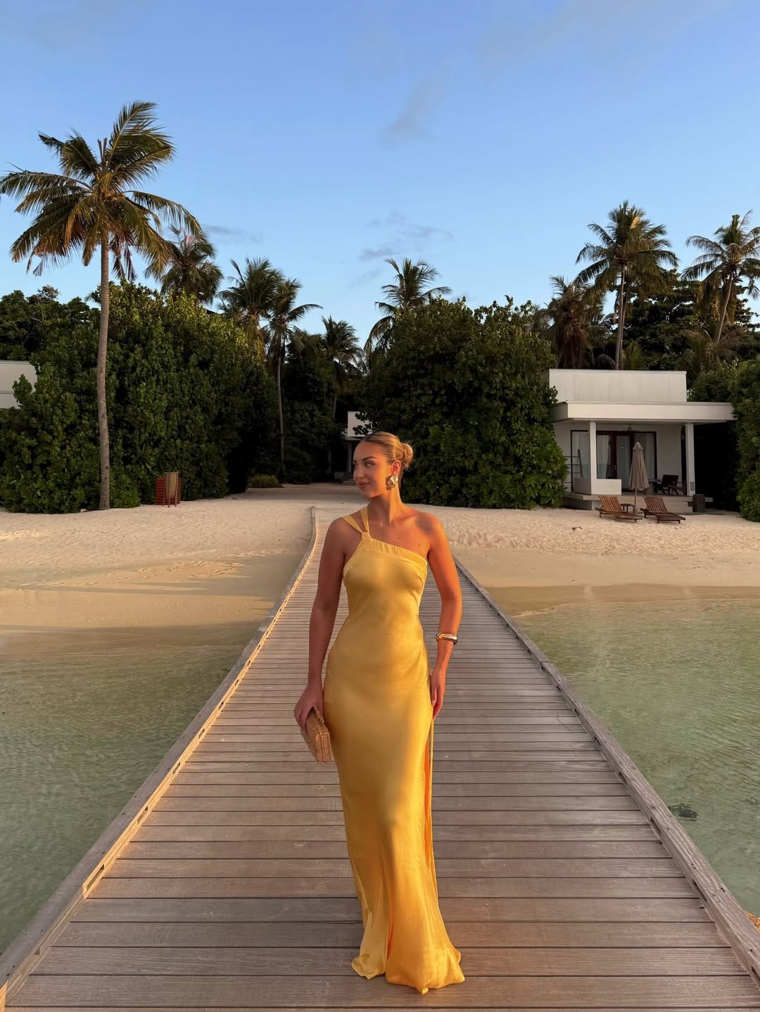 Woman in marigold yellow one-shoulder asymmetric strap column gown on an overwater boardwalk at sunset in the Maldives.