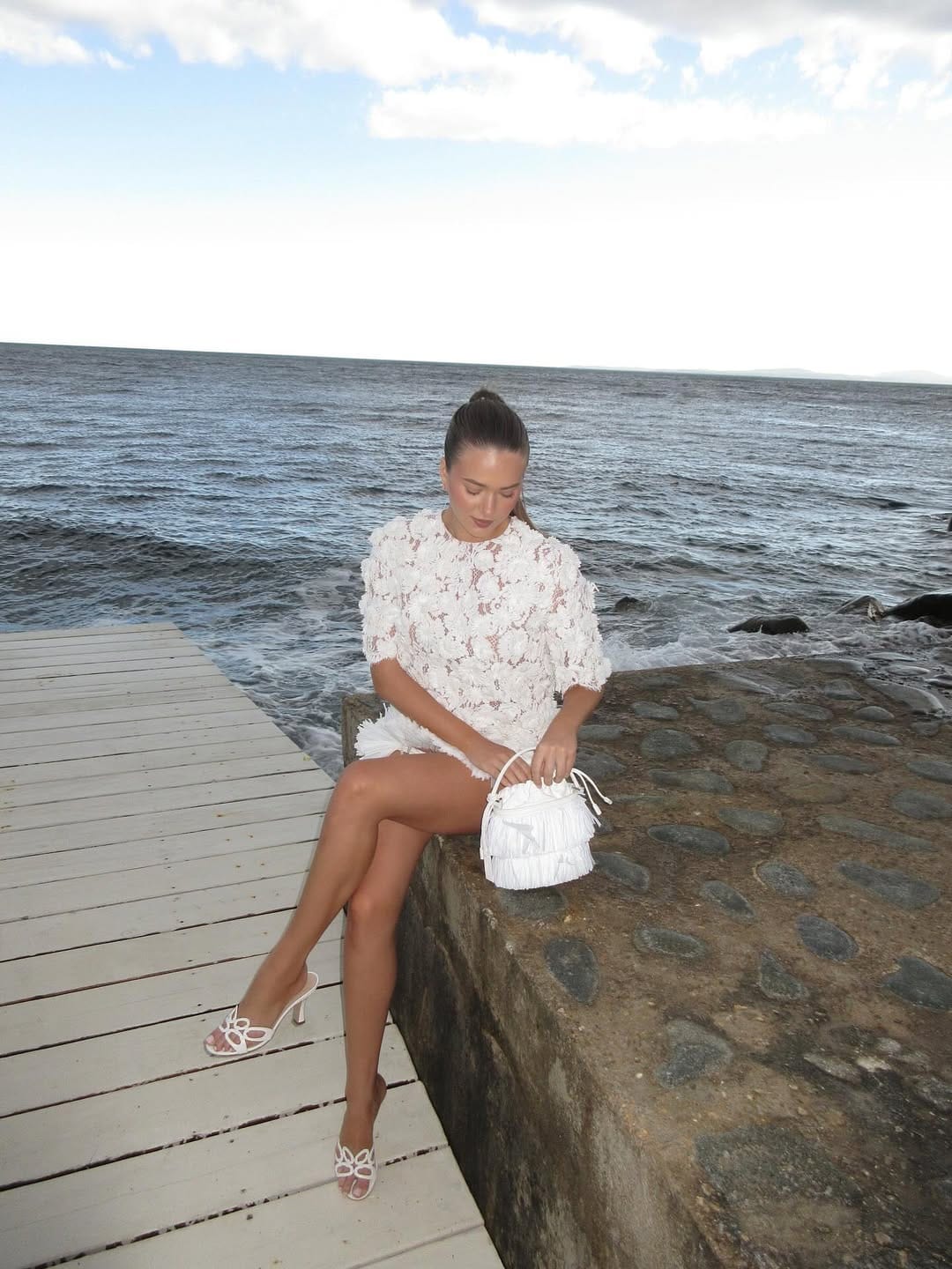 Woman in white 3D floral lace mini dress with bow heeled sandals and white fringe bucket bag by the sea.