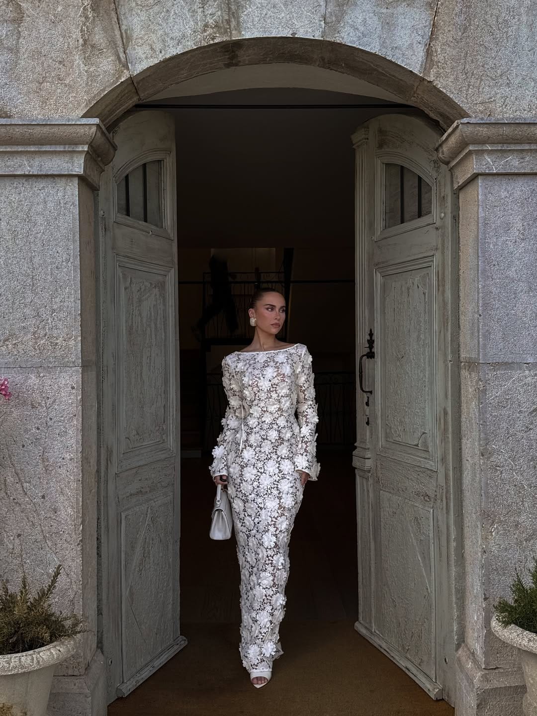 Woman in white long-sleeve 3D floral appliqué column gown with white bag in a stone doorway.