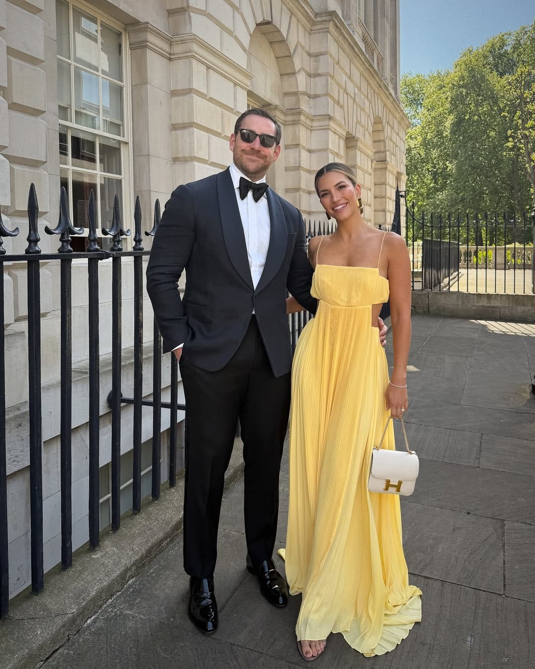 Woman in sunshine yellow pleated spaghetti-strap maxi gown with white Hermès bag outside a London building.