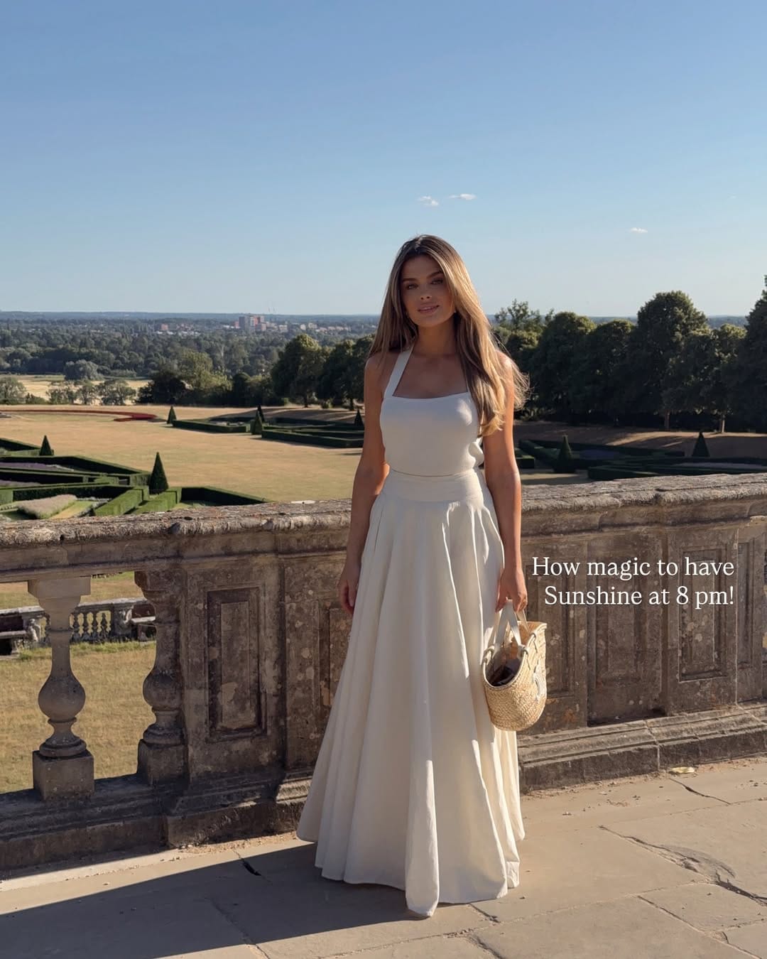 Woman in ivory halterneck full A-line maxi gown with woven basket bag on a grand stone terrace.