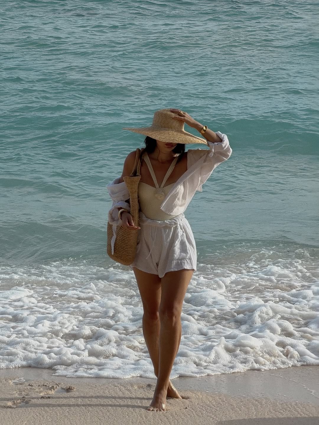 White linen shirt over cream swimsuit and shorts with straw tote and boater hat at shoreline beach outfit idea