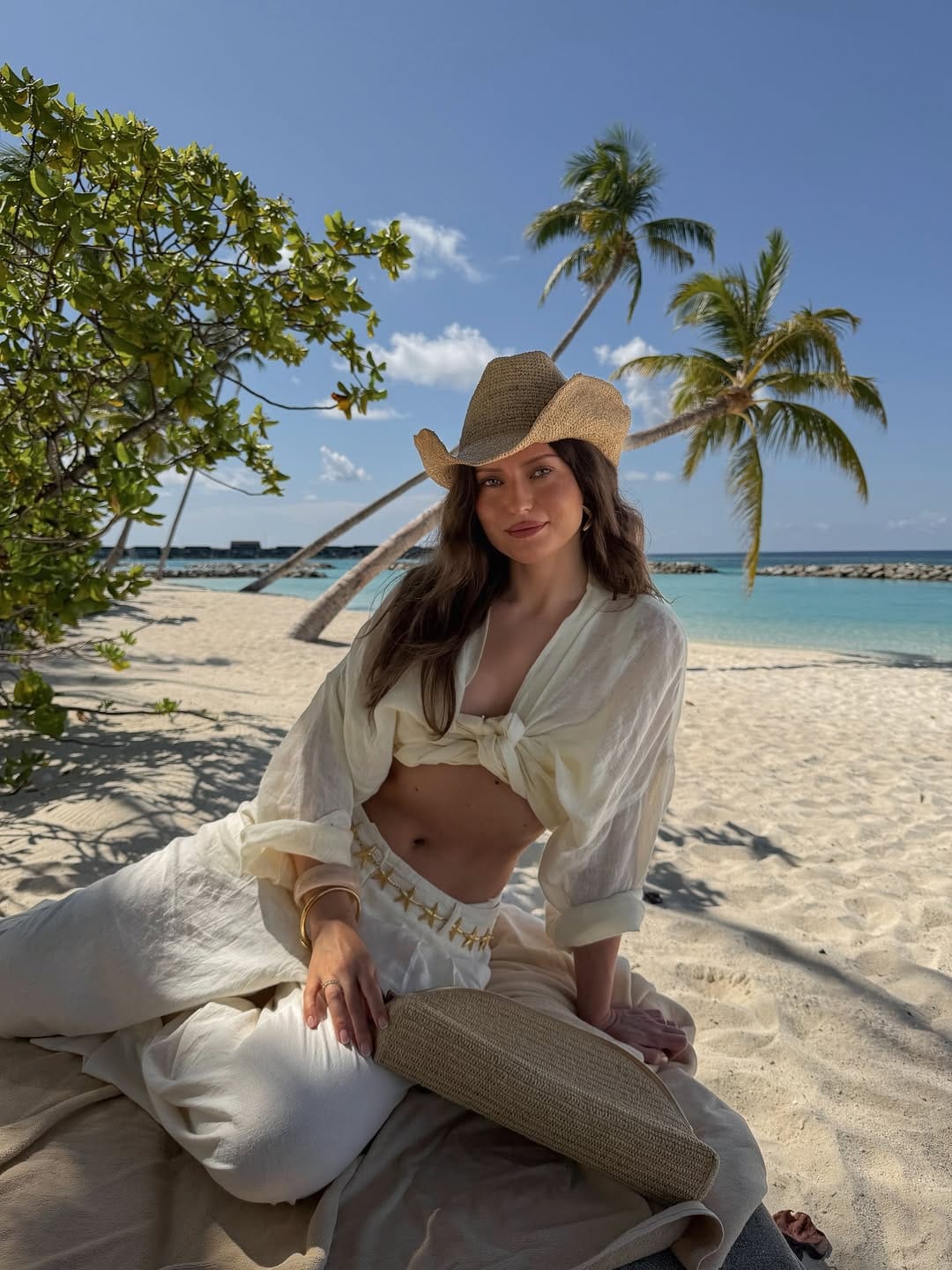 Cream tie-front linen shirt with white star-embroidered linen trousers and raffia cowboy hat on Maldives beach outfit idea