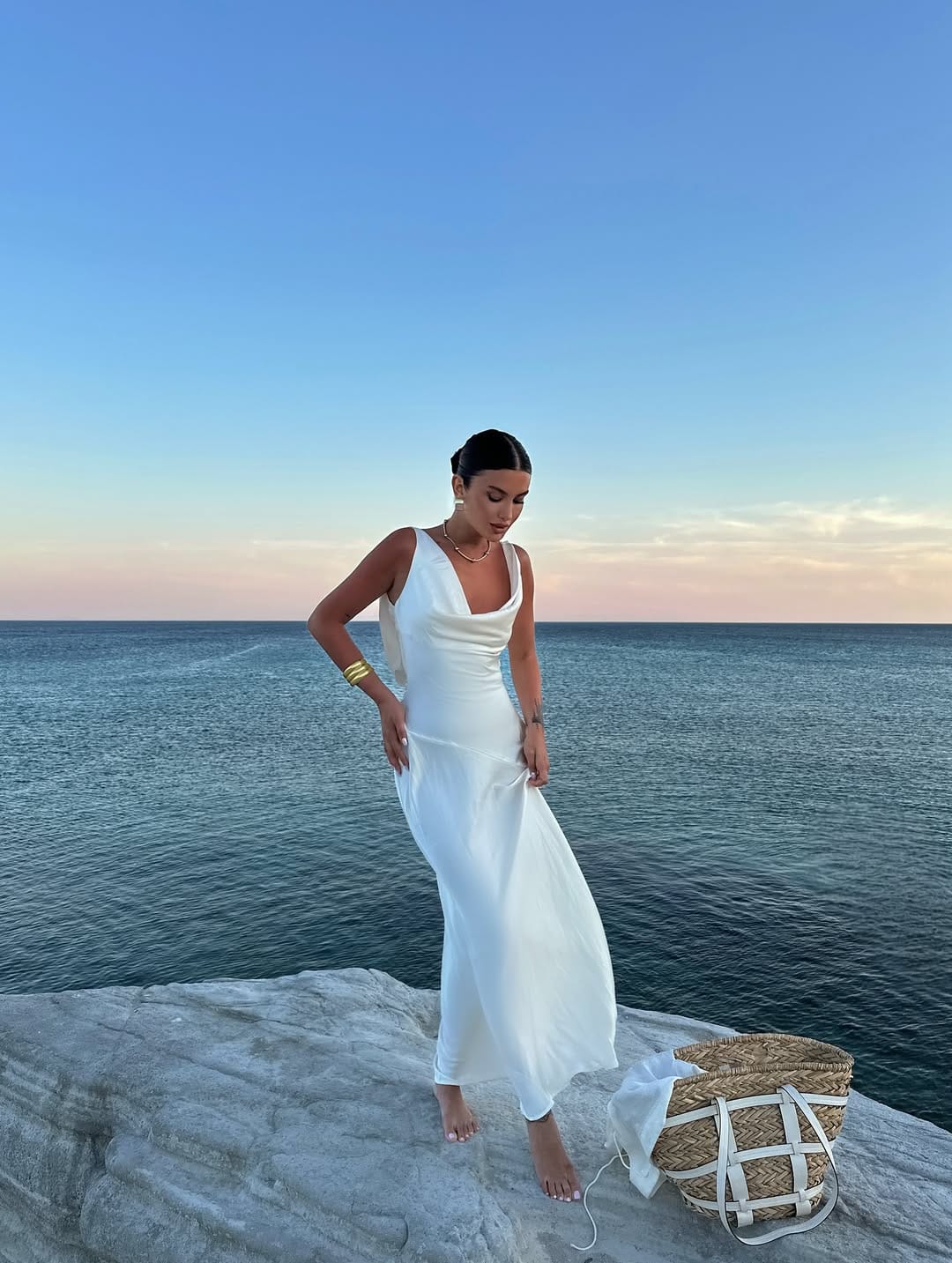 White satin cowl-neck slip maxi dress with gold cuff and straw tote on coastal rocks beach outfit idea