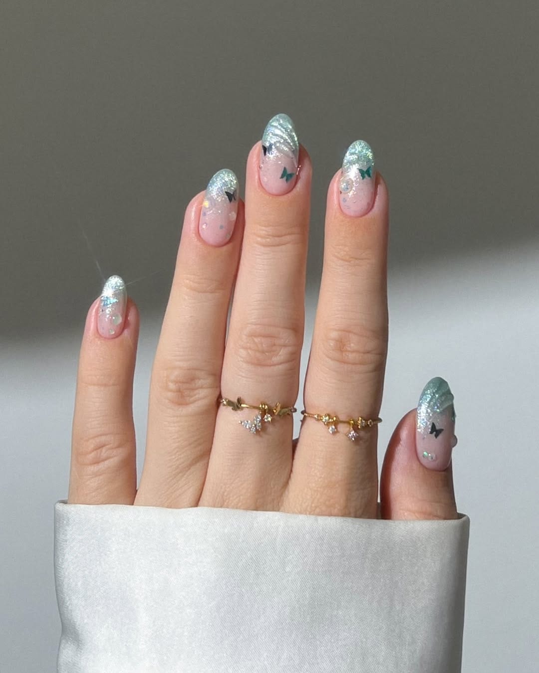 Sheer pink almond nails with silver glitter tips and micro butterfly accents