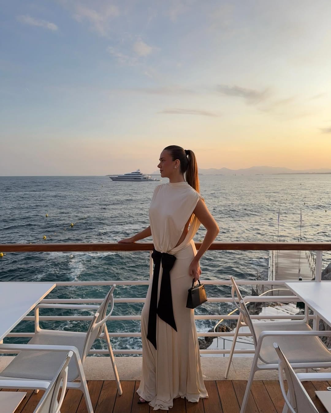 Cream draped jersey maxi dress with oversized black bow belt and small black bag at a seaside restaurant