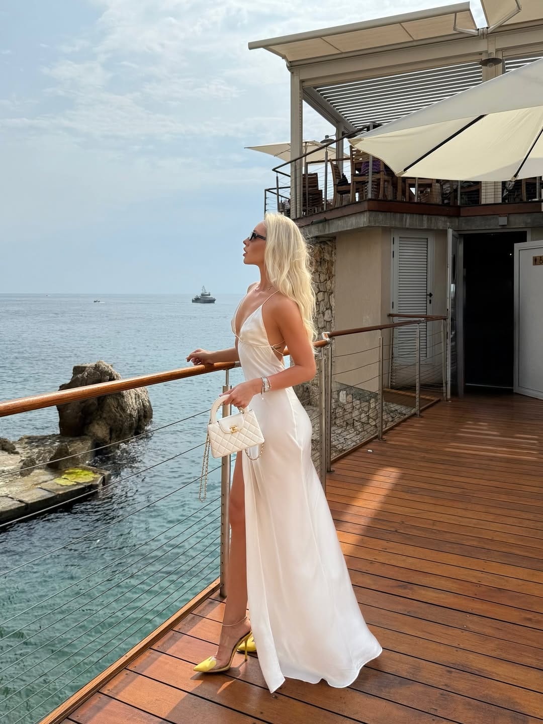 White satin slip dress with deep V-neckline and high slit with clear heels on a seaside terrace