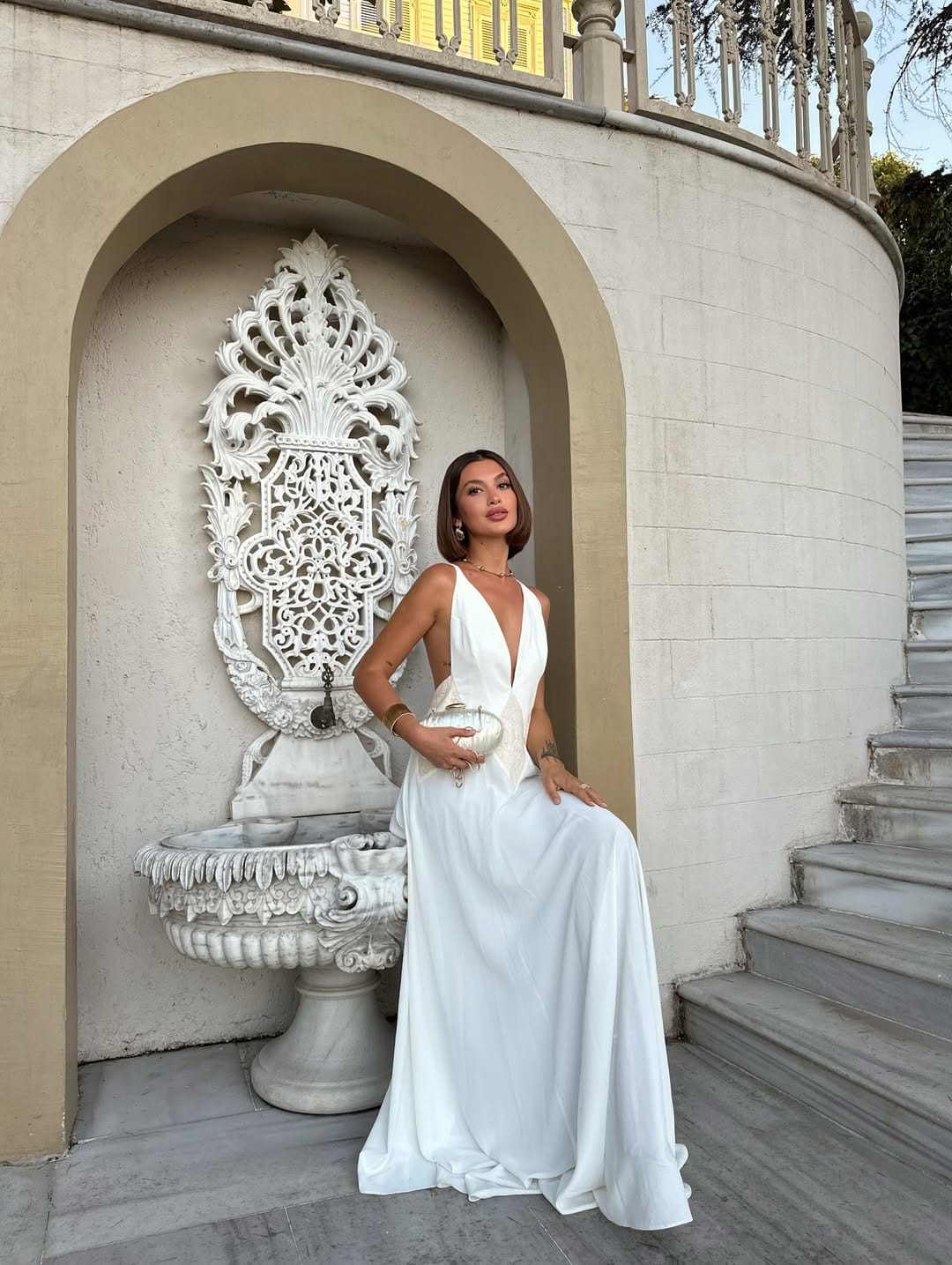 White deep-V satin gown with open back and silver clutch at a Mediterranean villa