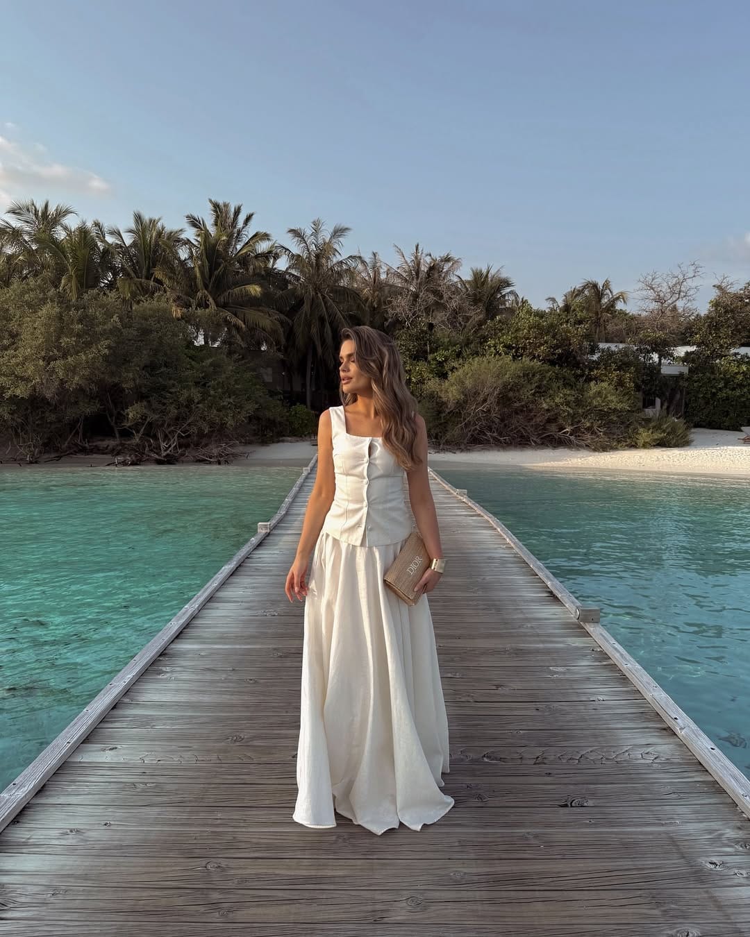 White structured corset top and voluminous linen maxi skirt with Dior book clutch on a tropical jetty