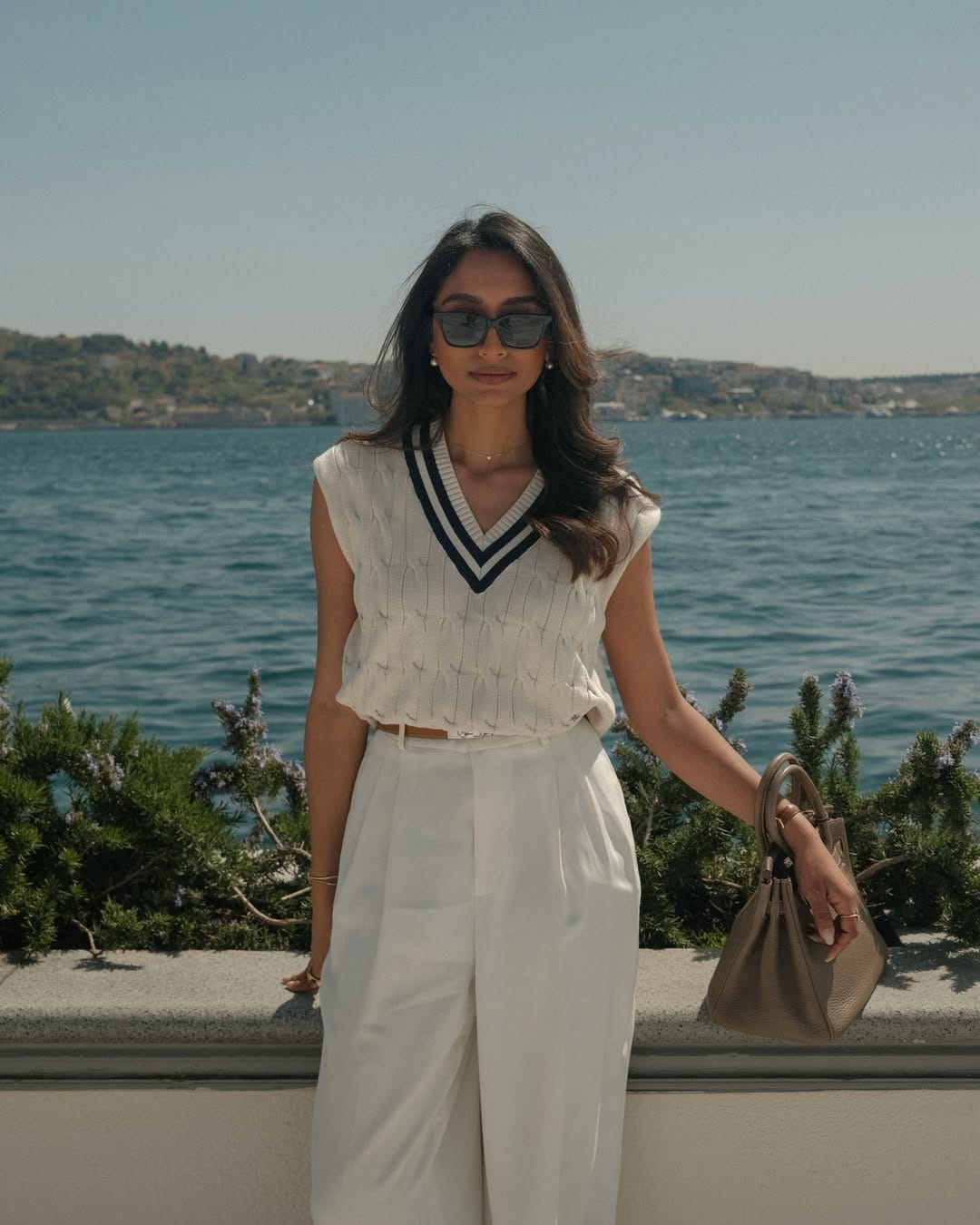 Cream cable-knit cricket vest with navy stripe trim and white wide-leg trousers by the sea