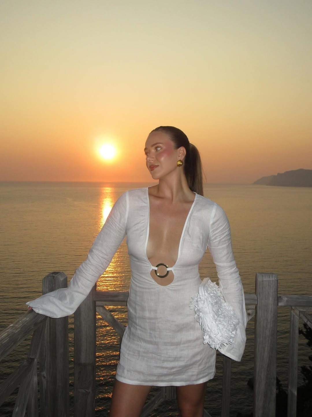 White linen deep-V mini dress with bell sleeves and gold earrings at Greece island sunset