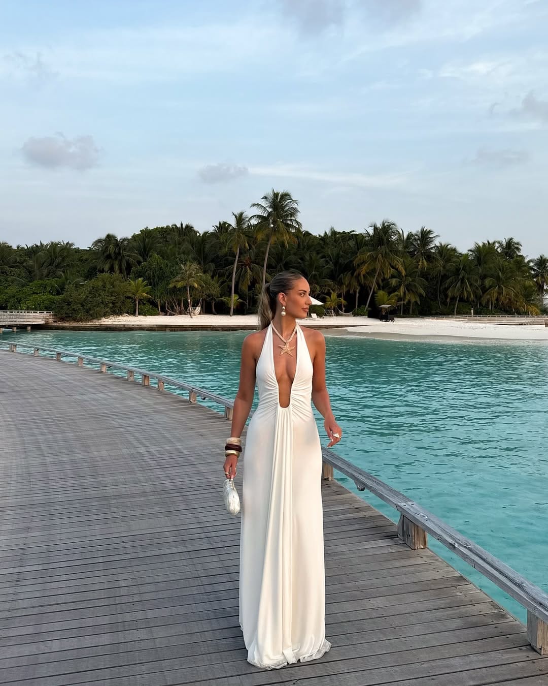 A white deep-plunge halter neck jersey maxi dress styled with pearl and starfish jewelry for a coastal-inspired look.
