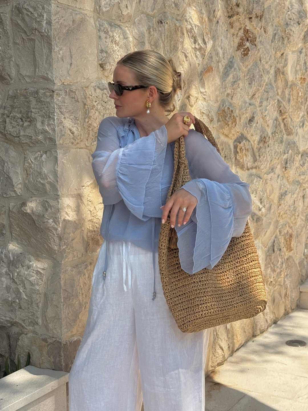 A powder blue sheer ruffle-sleeve blouse styled with white wide-leg linen trousers and a raffia tote for a light, airy outfit.