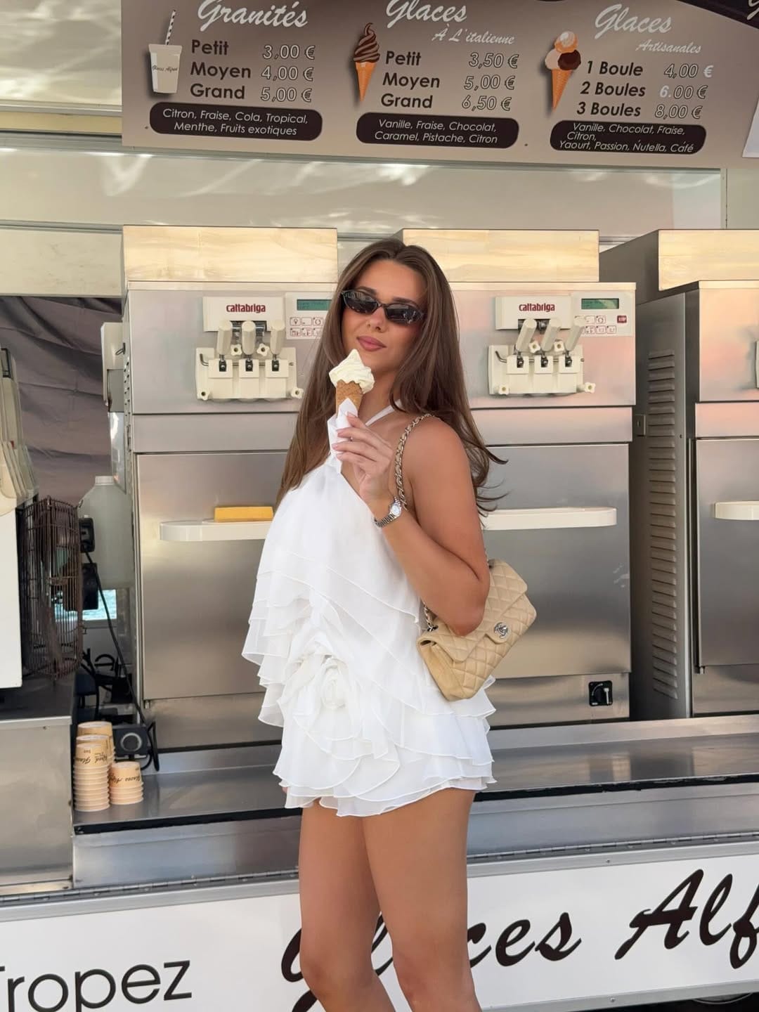 A white one-shoulder tiered ruffle mini dress styled with a beige quilted bag for a soft and feminine finish.