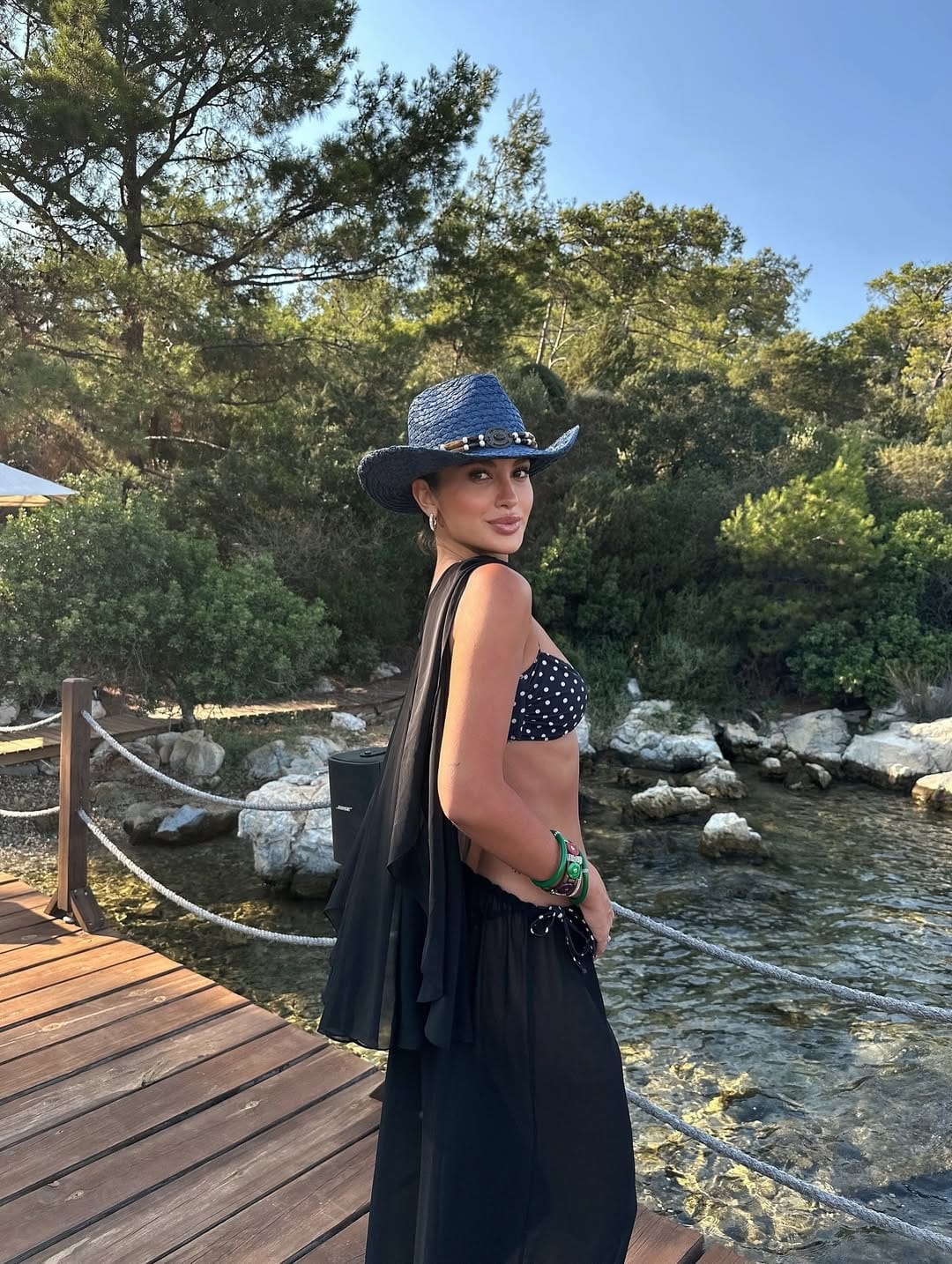 A navy polka dot bandeau bikini top styled with a sheer black chiffon sarong and a woven cowboy hat for a coastal vibe.