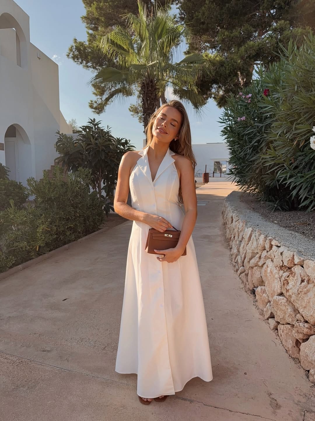 A white halter neck collared maxi dress paired with a tan mini bag, combining structure with effortless elegance.