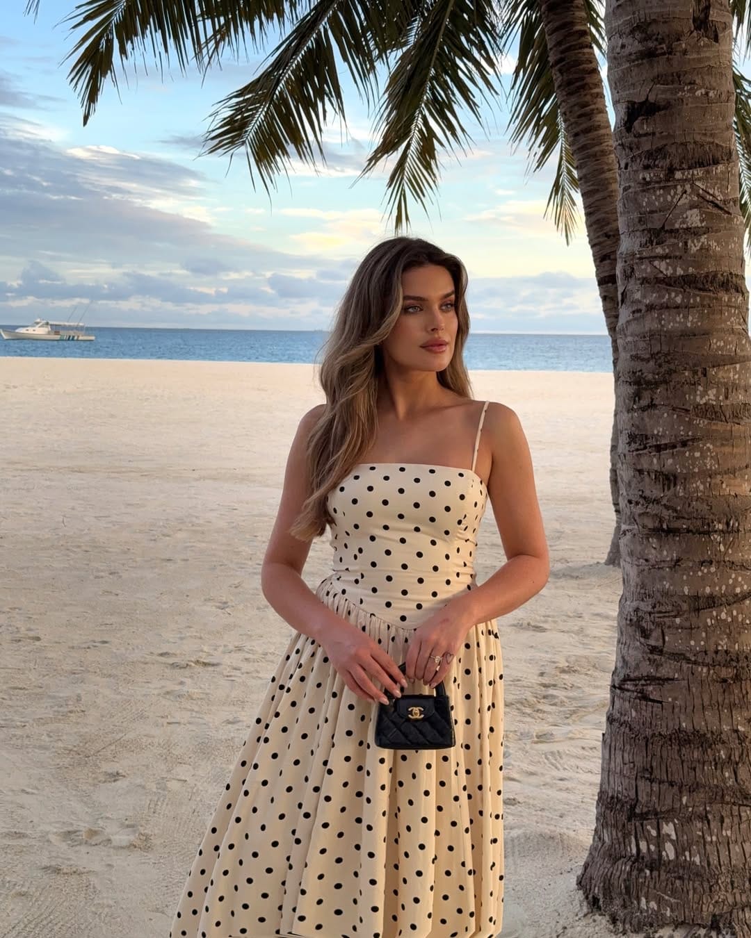 A cream polka dot strappy midi dress styled with a small quilted bag, perfect for a soft and romantic getaway look.