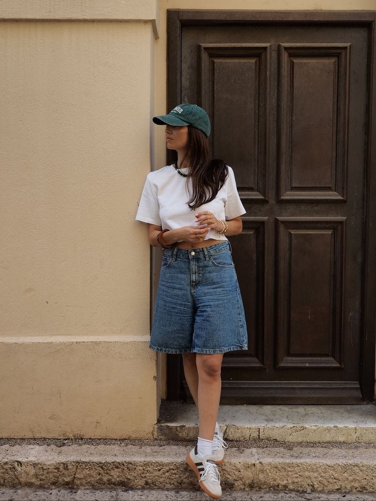 White cropped tee styled with a green cap, mid-length jorts, and Sambas for a sporty summer outfit.