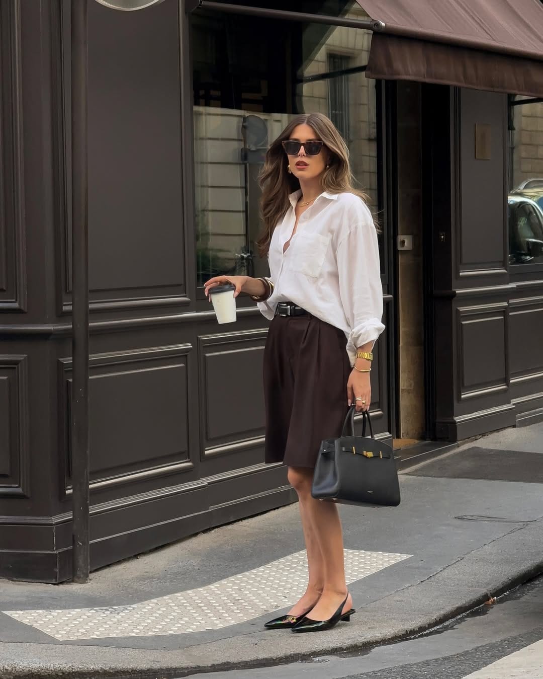  Woman in oversized white linen shirt and chocolate Bermuda shorts with black patent slingback heels, NYC summer outfit on a city street