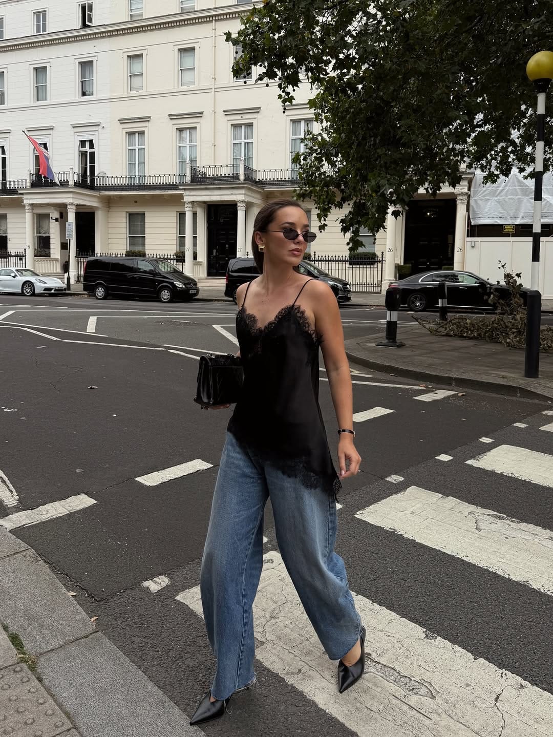  Woman in black lace slip cami and baggy wide-leg blue jeans with black kitten mules crossing a city street, summer street style New York