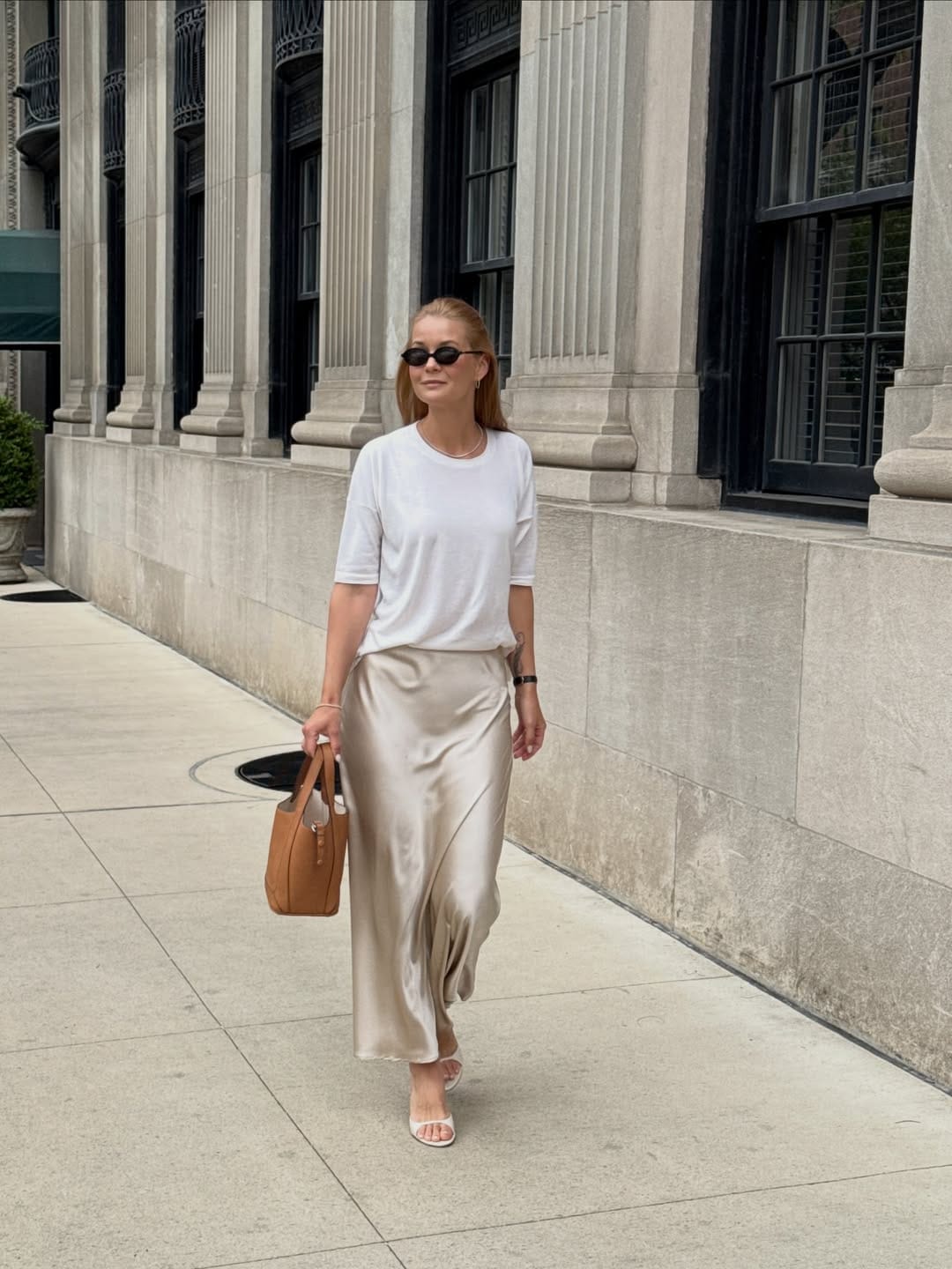 Woman in white crew-neck tee and champagne satin bias-cut maxi skirt with tan tote walking past NYC building columns, summer street style New York